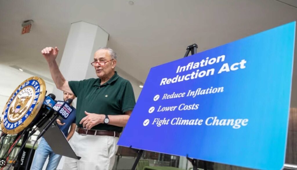 Senator Chuck Schumer standing behind a platform speaking while a poster explaining the Inflation Reduction Act sits in the foreground